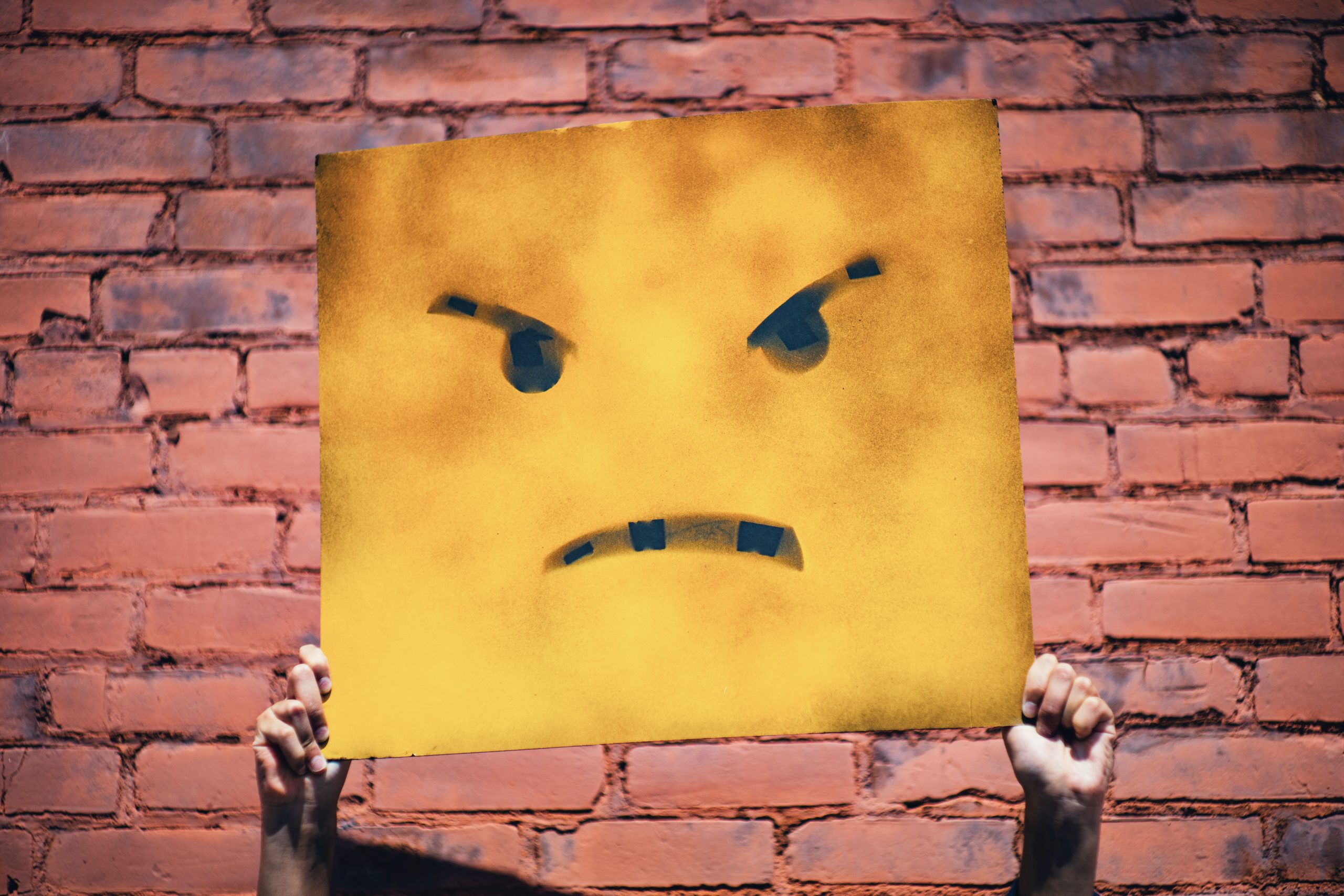 Dirty Yellow Box with a scowling face drawn on being held up in front of a persons face with a brick background